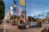 A town square with decorative banners displaying family names and crests mounted on poles. The area is surrounded by colorful flower arrangements and well-maintained trees, with modern benches and paved walkways. A metal sculpture and historic-looking buildings can be seen in the background, set against a sky transitioning from day to evening.