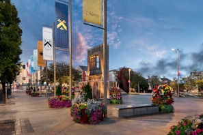 A town square with decorative banners displaying family names and crests mounted on poles. The area is surrounded by colorful flower arrangements and well-maintained trees, with modern benches and paved walkways. A metal sculpture and historic-looking buildings can be seen in the background, set against a sky transitioning from day to evening.
