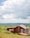 A small red cabin with a sloped roof is situated in a grassy field surrounded by lush greenery. Two propane tanks are placed outside the cabin near the wall. A red canoe rests on the ground beside the structure. The background features rolling hills under a partly cloudy sky, adding depth to the landscape.