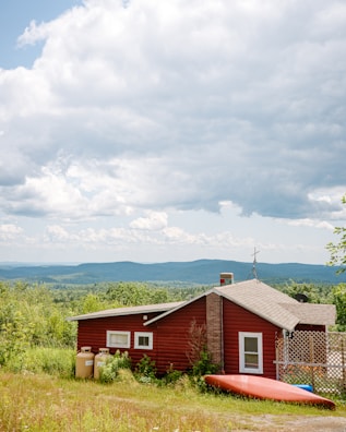 A small red cabin with a sloped roof is situated in a grassy field surrounded by lush greenery. Two propane tanks are placed outside the cabin near the wall. A red canoe rests on the ground beside the structure. The background features rolling hills under a partly cloudy sky, adding depth to the landscape.