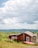 A small red cabin with a sloped roof is situated in a grassy field surrounded by lush greenery. Two propane tanks are placed outside the cabin near the wall. A red canoe rests on the ground beside the structure. The background features rolling hills under a partly cloudy sky, adding depth to the landscape.