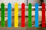 Colorful wooden fence slats painted in bright primary colors, including green, blue, yellow, and red, are arranged in an alternating pattern, mounted vertically and secured with metal screws. The background is a stone wall with a reflection visible on a wet concrete surface.