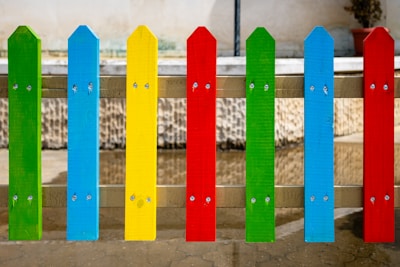 Colorful wooden fence slats painted in bright primary colors, including green, blue, yellow, and red, are arranged in an alternating pattern, mounted vertically and secured with metal screws. The background is a stone wall with a reflection visible on a wet concrete surface.