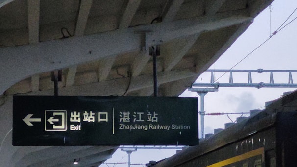A section of a railway station platform is visible, featuring a sign for 'ZhanJiang Railway Station' above an exit sign with directional arrows. Overhead wires and part of a train are also in view, beneath a large station roof.