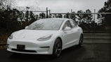 A sleek white electric car is parked on a wet asphalt surface in front of a chain-link fence topped with barbed wire. The car's modern design is highlighted against a backdrop of bare trees and overcast skies, suggesting a cold and gloomy day.