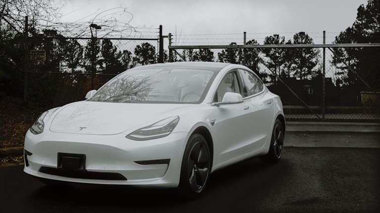 A sleek white electric car is parked on a wet asphalt surface in front of a chain-link fence topped with barbed wire. The car's modern design is highlighted against a backdrop of bare trees and overcast skies, suggesting a cold and gloomy day.
