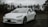 A sleek white electric car is parked on a wet asphalt surface in front of a chain-link fence topped with barbed wire. The car's modern design is highlighted against a backdrop of bare trees and overcast skies, suggesting a cold and gloomy day.