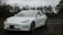 A sleek white electric car is parked on a wet asphalt surface in front of a chain-link fence topped with barbed wire. The car's modern design is highlighted against a backdrop of bare trees and overcast skies, suggesting a cold and gloomy day.