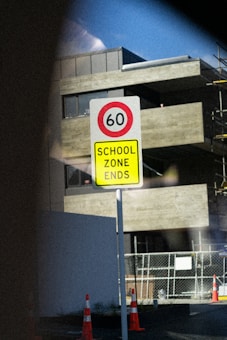 A road sign indicating the end of a school zone with a speed limit of 60, placed in an urban setting. The background features a modern building with large windows and a construction area fenced off with orange cones.