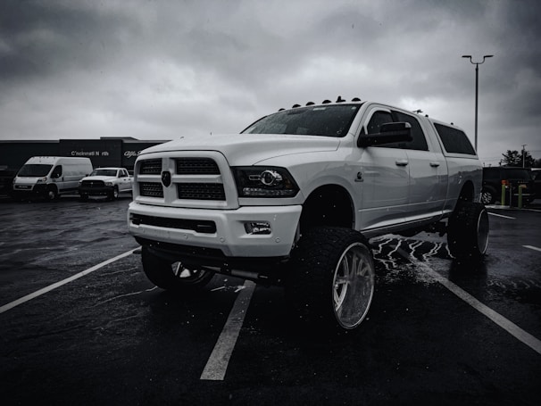 A large white pickup truck with lifted suspension and oversized tires is parked in a wet parking lot under overcast skies. Other vehicles, including vans, are visible in the background with a building bearing the signage of a business identifying the location as Cincinnati North.