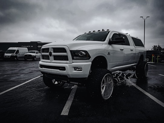 A large white pickup truck with lifted suspension and oversized tires is parked in a wet parking lot under overcast skies. Other vehicles, including vans, are visible in the background with a building bearing the signage of a business identifying the location as Cincinnati North.