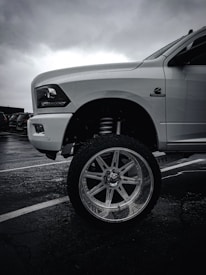 A high-contrast image featuring the front end of a large white pickup truck with an emphasis on its elevated suspension and oversized chrome wheel. The environment appears overcast, with dark, cloudy skies and a wet parking lot surface.