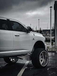 A white pickup truck with large, custom wheels is parked in a lot on a cloudy day. The truck features elevated suspension and is prominently set against a backdrop of overcast skies and urban surroundings, including street lights and a sign.