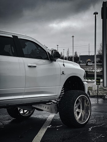 A white pickup truck with large, custom wheels is parked in a lot on a cloudy day. The truck features elevated suspension and is prominently set against a backdrop of overcast skies and urban surroundings, including street lights and a sign.