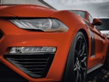 Close-up of a sleek, orange sports car's front, highlighting its shiny bodywork and intricate design details. The car's headlight is prominently featured, alongside a wet surface from rain droplets, adding a dynamic and fresh appearance.