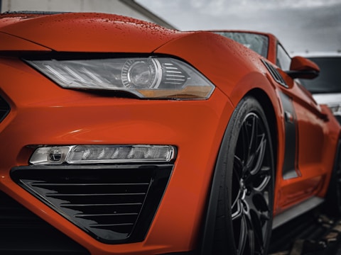 Close-up of a sleek, orange sports car's front, highlighting its shiny bodywork and intricate design details. The car's headlight is prominently featured, alongside a wet surface from rain droplets, adding a dynamic and fresh appearance.