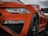 Close-up of a sleek, orange sports car's front, highlighting its shiny bodywork and intricate design details. The car's headlight is prominently featured, alongside a wet surface from rain droplets, adding a dynamic and fresh appearance.
