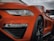 Close-up of a sleek, orange sports car's front, highlighting its shiny bodywork and intricate design details. The car's headlight is prominently featured, alongside a wet surface from rain droplets, adding a dynamic and fresh appearance.