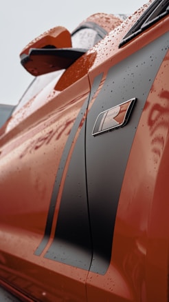 Close-up view of the side of an orange car featuring a sleek black racing stripe and an 'R' emblem. The surface appears wet, suggesting recent rain, with droplets glistening on the body.