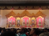 A stage with ornate golden decorations and vibrant pink backdrops features religious figures adorned with elaborate clothing and jewelry. Framed portraits of spiritual leaders are displayed between the figures. The foreground shows a large crowd of people observing the scene.