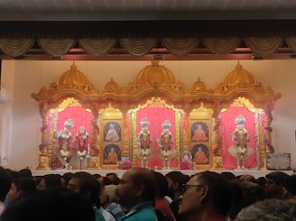 A stage with ornate golden decorations and vibrant pink backdrops features religious figures adorned with elaborate clothing and jewelry. Framed portraits of spiritual leaders are displayed between the figures. The foreground shows a large crowd of people observing the scene.