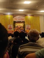 An indoor gathering with people seated, facing a decorated stage with colorful flower garlands. The stage displays portraits and a small altar with offerings, indicating a cultural or religious event. Participants are dressed casually. Fluorescent lights illuminate the room from the ceiling with visible fans.