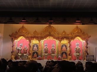 An ornate altar with golden decorative elements houses several statues dressed in colorful, traditional garments. The background features portraits of revered figures, framed by rich red drapes and intricate gold accents. The setup is lavishly adorned with floral garlands, indicating a religious or ceremonial setting. In the foreground, a crowd of people observes the altar.