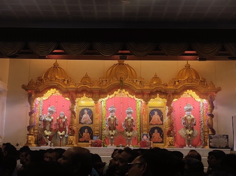 An ornate altar with golden decorative elements houses several statues dressed in colorful, traditional garments. The background features portraits of revered figures, framed by rich red drapes and intricate gold accents. The setup is lavishly adorned with floral garlands, indicating a religious or ceremonial setting. In the foreground, a crowd of people observes the altar.