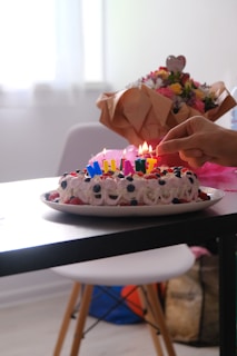 A decorated cake with colorful candles is on a table, surrounded by fruits like blueberries and strawberries. A hand is seen lighting the candles, and there is a bouquet of flowers wrapped in brown paper in the background.