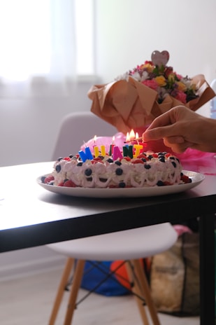 A decorated cake with colorful candles is on a table, surrounded by fruits like blueberries and strawberries. A hand is seen lighting the candles, and there is a bouquet of flowers wrapped in brown paper in the background.