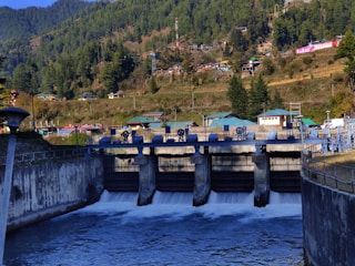 A dam with multiple sluice gates releases water into a river. Green forests cover the hills in the background, with several small houses scattered along the hillside. The sky above is clear, and the terrain appears to be a mixture of grass and rocky pathways.