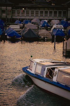 A small boat with a blue roof moves through calm water, leaving a gentle wake behind. Covered boats are docked in the background, protected by tarps in shades of blue and gray. The surrounding architecture exhibits large windows and a dimly lit atmosphere, suggesting evening or early morning.