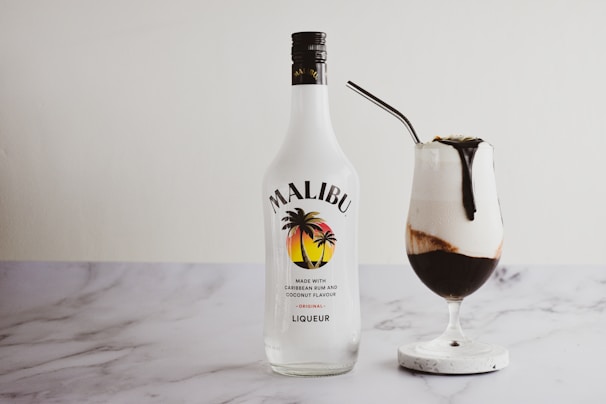 A bottle of Malibu, a Caribbean rum with coconut flavor, is placed next to a cocktail in a glass. The cocktail appears creamy with a dark chocolate or coffee topping, and is served with a straw on a marble coaster.