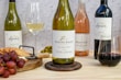 A selection of wine bottles is displayed on a wooden table featuring a Kleine Zalze Sauvignon Blanc 2022, a Spier Chenin Blanc, a Warwick The First Lady Rosé, and a Spier Merlot. The setup includes a platter with red grapes, crackers, and green olives next to a cheese plane. Two wine glasses, one filled with white wine and the other with red wine, accompany the arrangement against a speckled background.