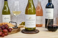 A selection of wine bottles is displayed on a wooden table featuring a Kleine Zalze Sauvignon Blanc 2022, a Spier Chenin Blanc, a Warwick The First Lady Rosé, and a Spier Merlot. The setup includes a platter with red grapes, crackers, and green olives next to a cheese plane. Two wine glasses, one filled with white wine and the other with red wine, accompany the arrangement against a speckled background.