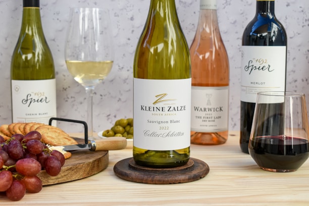 A selection of wine bottles is displayed on a wooden table featuring a Kleine Zalze Sauvignon Blanc 2022, a Spier Chenin Blanc, a Warwick The First Lady Rosé, and a Spier Merlot. The setup includes a platter with red grapes, crackers, and green olives next to a cheese plane. Two wine glasses, one filled with white wine and the other with red wine, accompany the arrangement against a speckled background.
