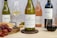 A selection of wine bottles is displayed on a wooden table featuring a Kleine Zalze Sauvignon Blanc 2022, a Spier Chenin Blanc, a Warwick The First Lady Rosé, and a Spier Merlot. The setup includes a platter with red grapes, crackers, and green olives next to a cheese plane. Two wine glasses, one filled with white wine and the other with red wine, accompany the arrangement against a speckled background.