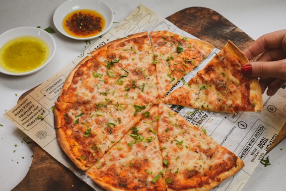 A freshly baked thin-crust pizza is sliced and topped with cheese and herbs. A hand with red nail polish is lifting one slice. Two small bowls of dipping sauces, one with a yellowish oil and another with a darker sauce, are placed nearby. The pizza rests on parchment paper with printed text, sitting on a wooden serving board.