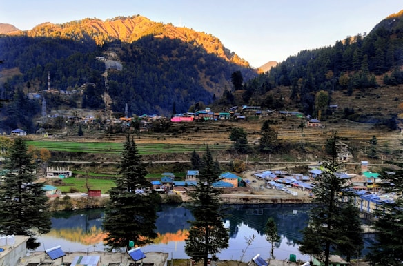 A serene mountainous landscape featuring a quaint village nestled among forested hills. The sunlight casts a warm glow on the peaks, and a calm river reflects the vibrant colors. The area is dotted with small houses and fields, surrounded by dense coniferous trees.