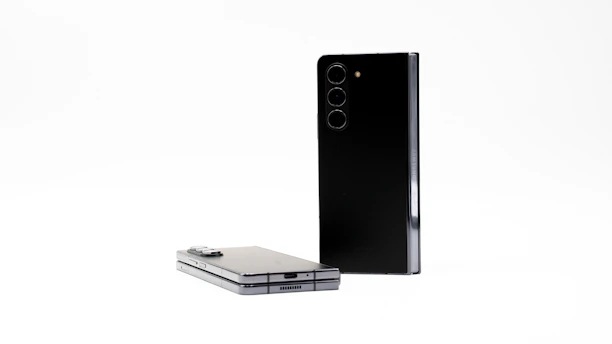 Two sleek, modern smartphones are displayed against a white background. One phone is folded closed and lies flat, showcasing its sides, which include buttons and ports. The other phone is upright, revealing the back with three camera lenses and a flash.