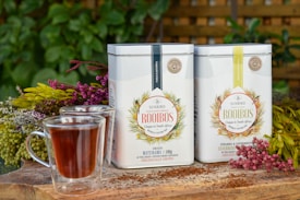 Two tins of Sunbird Rooibos tea are placed on a wooden surface surrounded by colorful flowers. There are two glasses of rooibos tea in the foreground, and lush green foliage is visible in the background. The scene is set outdoors, giving a fresh and natural vibe.