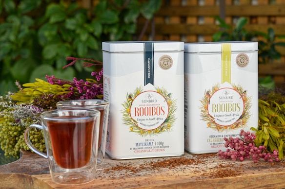 Two tins of Sunbird Rooibos tea are placed on a wooden surface surrounded by colorful flowers. There are two glasses of rooibos tea in the foreground, and lush green foliage is visible in the background. The scene is set outdoors, giving a fresh and natural vibe.