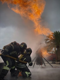Firefighters in protective gear are actively combating a large fire, with flames and smoke billowing into the sky. They are using a hose to spray water on the fire, and their determined posture shows intense focus on controlling the blaze. Palm trees are visible in the background, suggesting an outdoor environment.