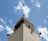 A tall beige and dark gray tower features multiple loudspeakers mounted at different angles. The sky is bright blue with scattered white clouds, offering a striking contrast to the structure's colors. At the pinnacle of the tower is an ornamental finial.