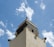 A tall beige and dark gray tower features multiple loudspeakers mounted at different angles. The sky is bright blue with scattered white clouds, offering a striking contrast to the structure's colors. At the pinnacle of the tower is an ornamental finial.