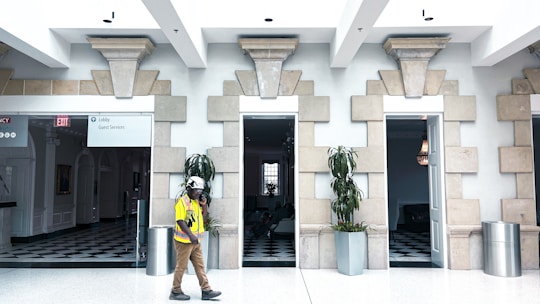 A person wearing a hard hat and a yellow safety vest walks past a building interior with stone architectural details. There are two open doorways with potted plants beside them, and a checkered black-and-white floor can be seen inside. One doorway leads to what appears to be a lobby or guest services area.