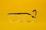 Clear eyeglasses with black arms are centered against a bright yellow background. The glasses feature a small, discreet logo on one lens and text on the arm, including 'Made in China' and model details.