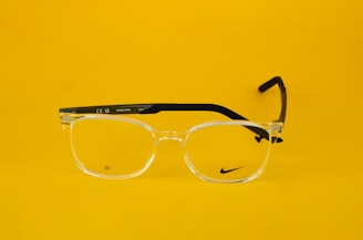 Clear eyeglasses with black arms are centered against a bright yellow background. The glasses feature a small, discreet logo on one lens and text on the arm, including 'Made in China' and model details.