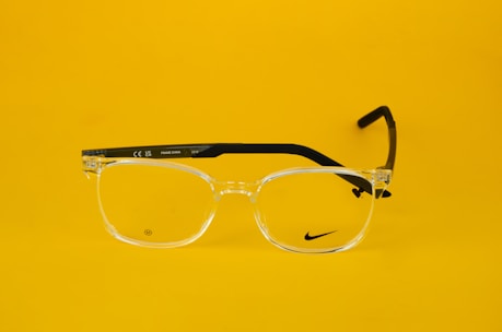 Clear eyeglasses with black arms are centered against a bright yellow background. The glasses feature a small, discreet logo on one lens and text on the arm, including 'Made in China' and model details.