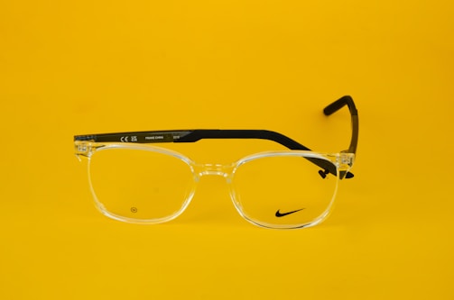 Clear eyeglasses with black arms are centered against a bright yellow background. The glasses feature a small, discreet logo on one lens and text on the arm, including 'Made in China' and model details.